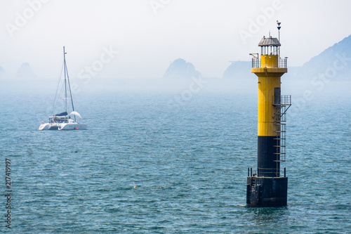 Yellow black lighthouse standing in the sea. There are several seagulls at the top. And small catamaran sail boat anchor near by.