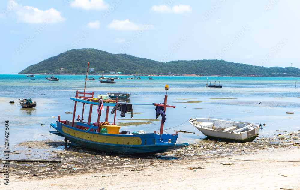 Small wooden and fiberglass fishing boats mooring at the dirty beach in front of fisherman village in Thailand on the sunny day with big island in the background