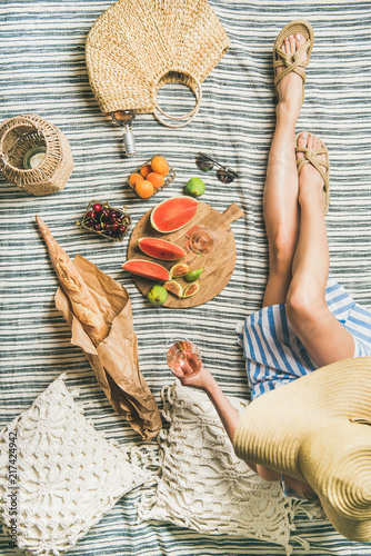 Summer picnic setting. Young woman in striped dress and straw sunhat sitting with glass of rose wine in hand, fresh fruit on board and baguette on blanket, top view. Outdoor gathering or lunch concept