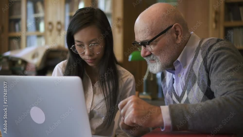 biologist Professor and female student are talking at table with laptop in library, elderly man and young woman are discussing scientific topic, sitting at desk with PC in reading room. Concept