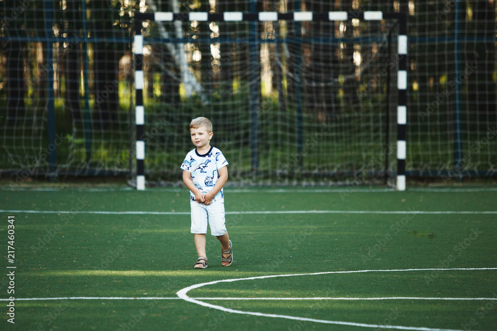 Happy cute blond boy in blue t-shirt and shorts on a football field without a ball.