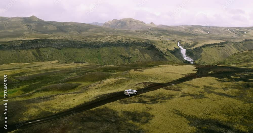 Aerial shot of 4x4 SUV car driving on top of mountain road with waterfall in background 