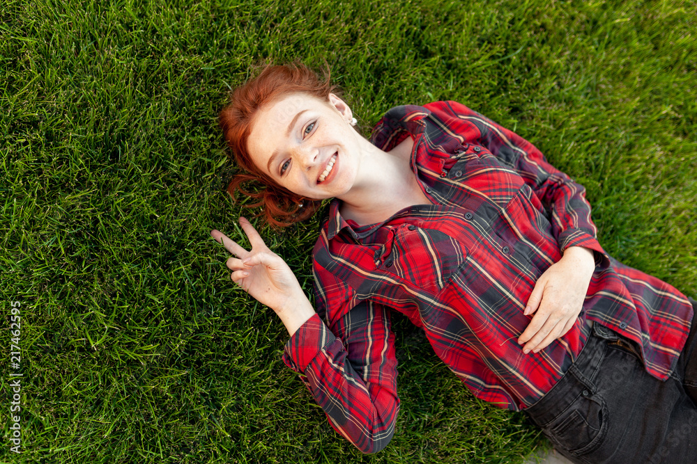 Beautiful young red-haired student girl with freckles lying on her back on grass and lawn view from above. Dressed in a red checkered shirt, resting after classes Beautiful young red-haired student girl with freckles lying on her back on grass and lawn view from above. Dressed in a red checkered shirt, resting after classes