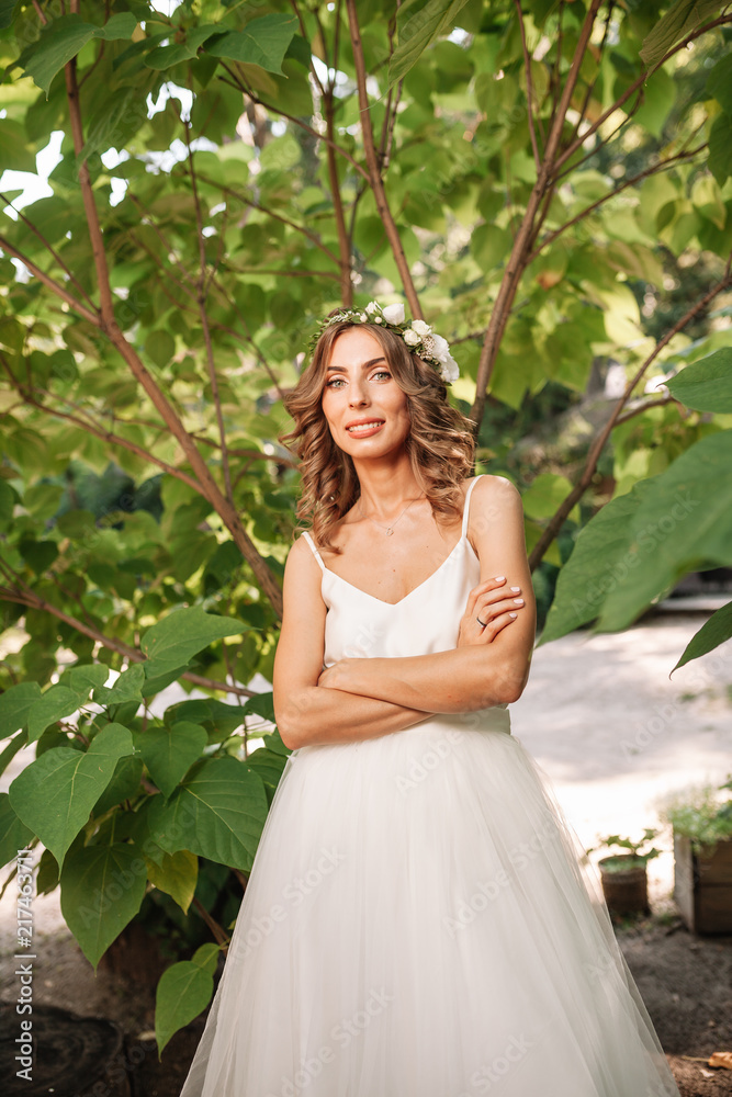 beautiful girl in white dress with a wreath of flowers on her head is standing outdoor in the park with green leaf on the background