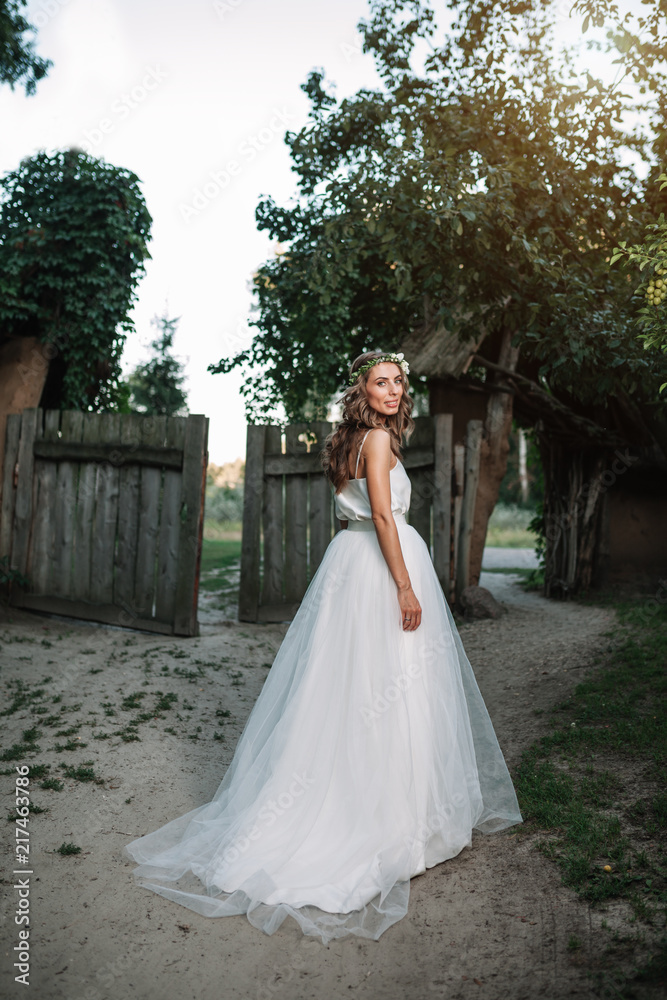 Fototapeta premium A cute curly woman in a white wedding dress with a wedding bouquet and wreath in her hair standing back to the camera in nature. Concept escaped bride. Forward to a happy bright future Runaway