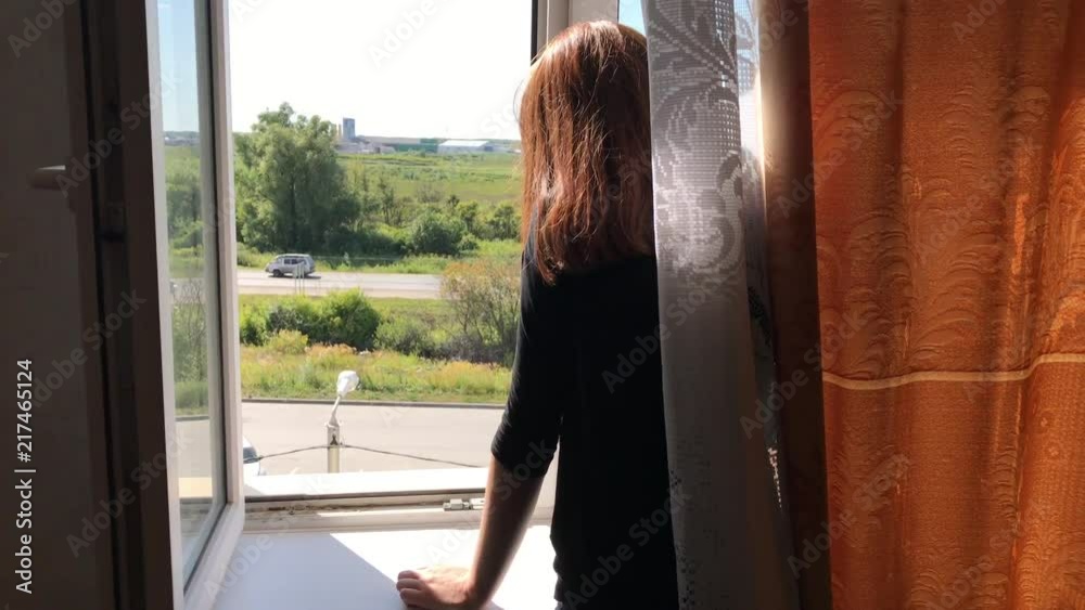 woman stands near an open window in the apartment and looks at the passing cars on the road woman stands near an open window in the apartment and looks at the passing cars on the road