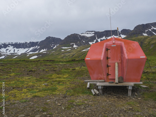 Red emergency shelter cabin in hloduvik campsite standing on the grass meadow with view on snow coved mountain cliffs, Hornstrandir, west fjords, Iceland photo