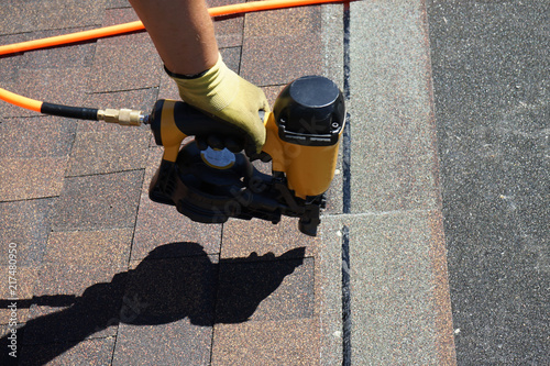 Roofer builder worker with nailgun installing Asphalt Shingles or Bitumen Tiles on a roof of a new house.