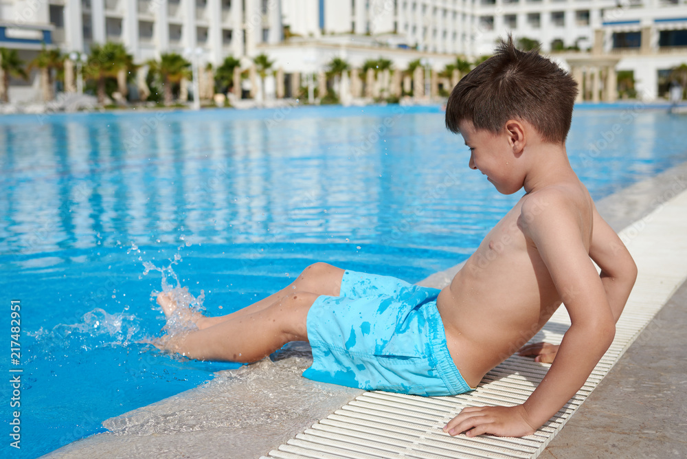 Smiling Caucasian boy having fun in pool at resort. He is holding legs in water.