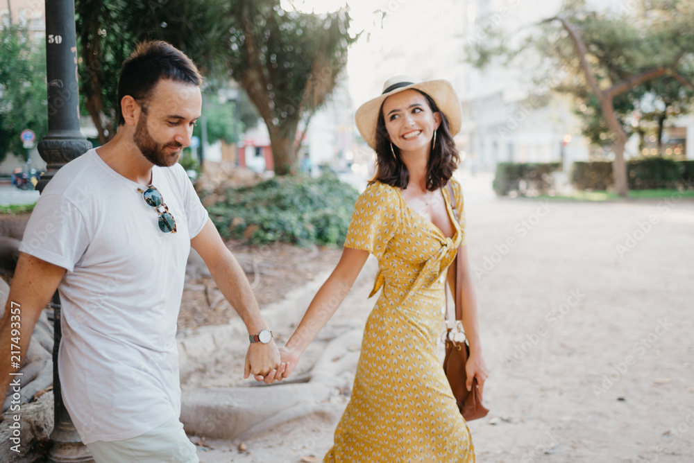 Beautiful young brunette woman with her boyfriend with beard walking holding hands under the giant tree in the city park in Spain in the evening. Intimate photo