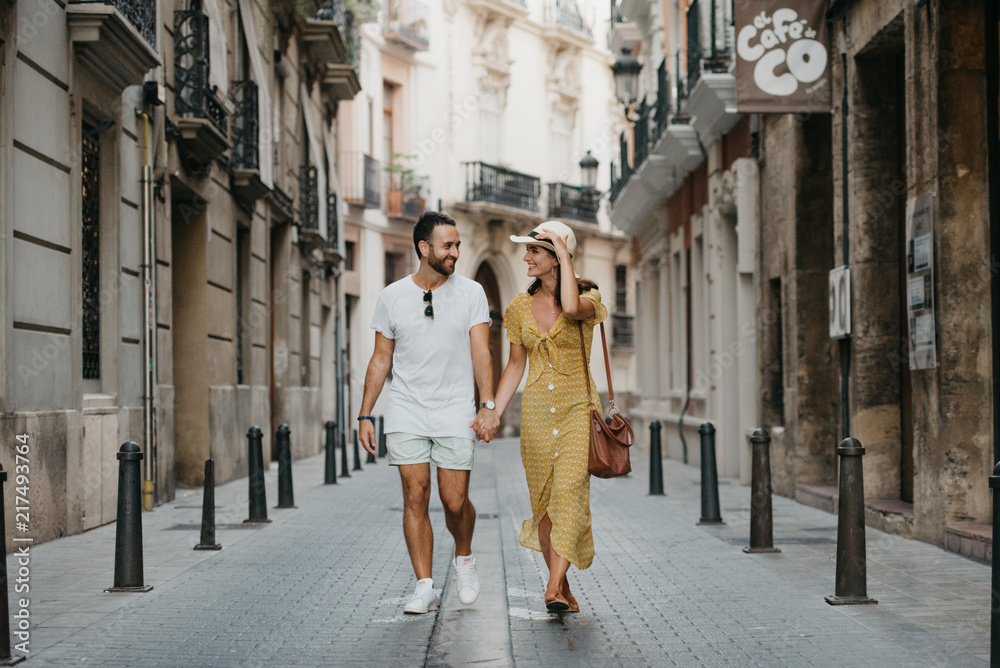Stylish brunette girl in the hat with her boyfriend with beard walking together in the center of the old European street in Spain in the evening