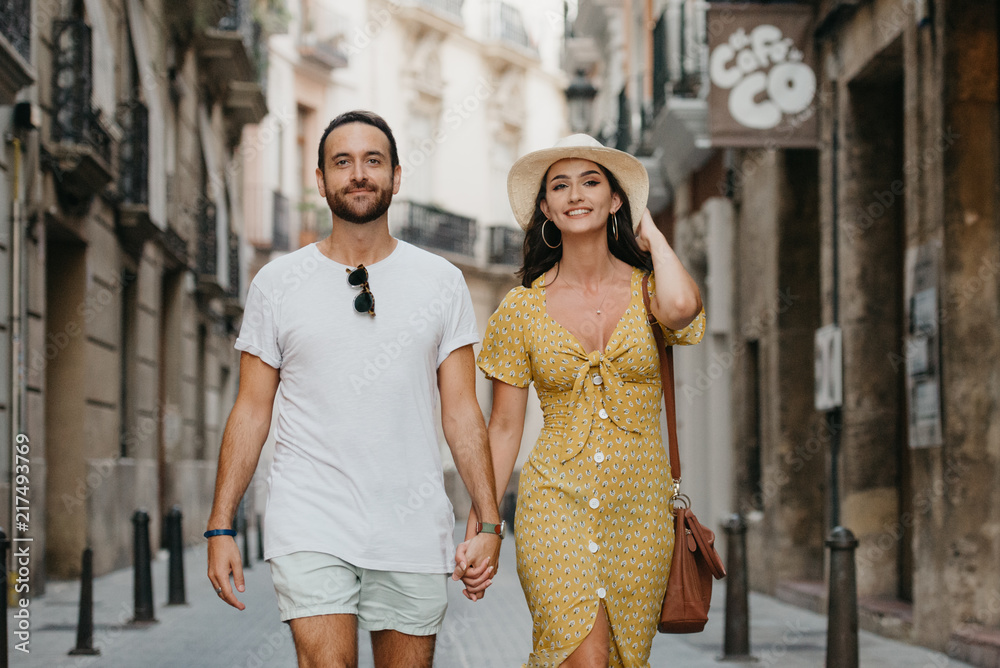 Stylish brunette girl in the hat with her boyfriend with beard walking together in the center of the old European street in Spain in the evening. Close portrait 