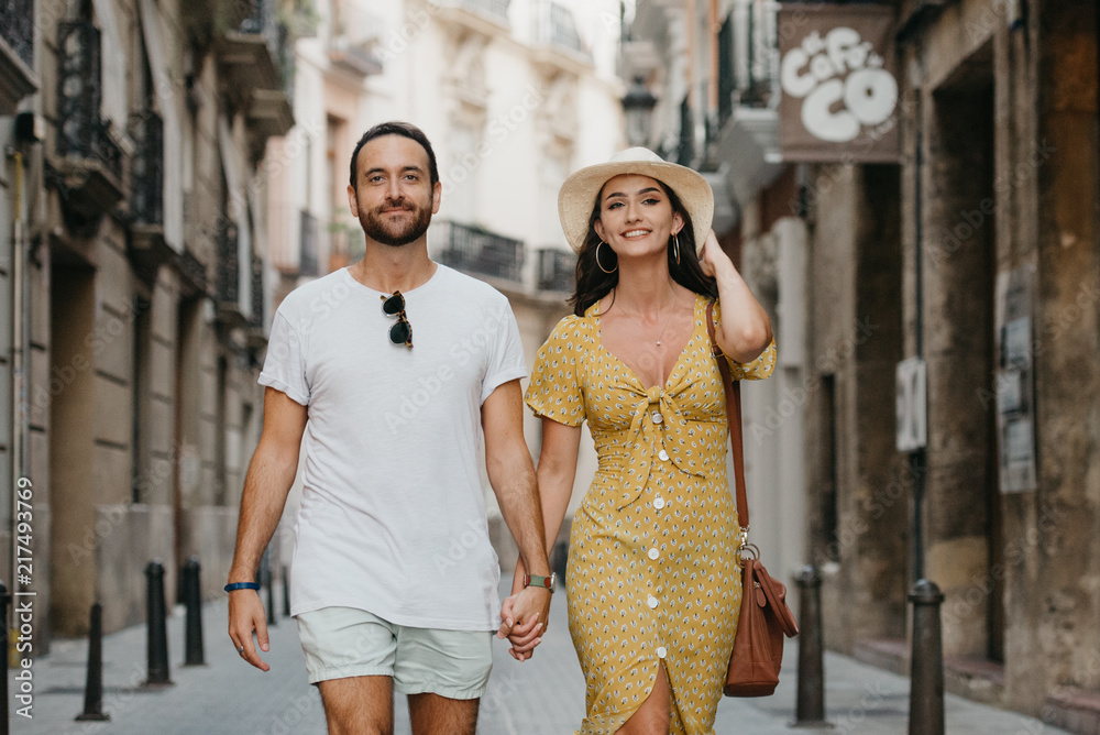 Stylish brunette girl in the hat with her boyfriend with beard walking together in the center of the old European street in Spain in the evening. Close portrait