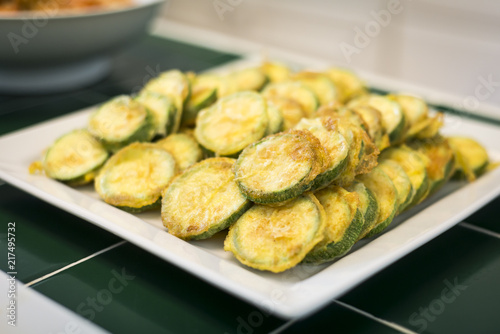 Korean pan fried zucchini on a white ceramic plate.  Resting on a green tile counter top.  Bowl of soup in the background.