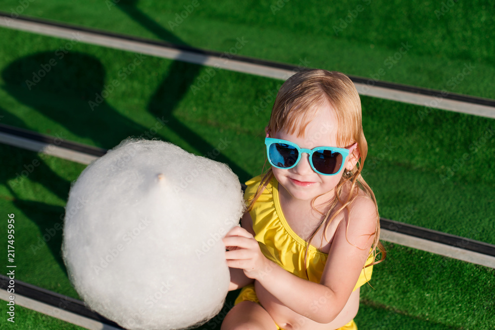 Beautiful 5 years old girl with blue sunglasses and wet hair, wearing in  yellow swimsuit eating sweet cotton wool, or cotton candy, or candy floss  on green background. foto de Stock