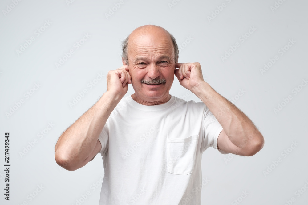 Mature man in white tshirt suffering from loud music and pain screaming trying to plug ears against grey background. Mature man in white tshirt suffering from loud music and pain screaming trying to plug ears against grey background.