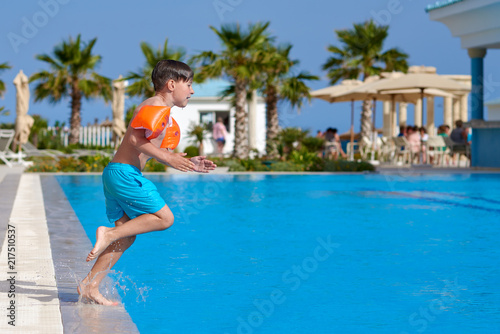 European boy in floating sleeves having fun jumping into swimming pool at resort. Side view. © Artem