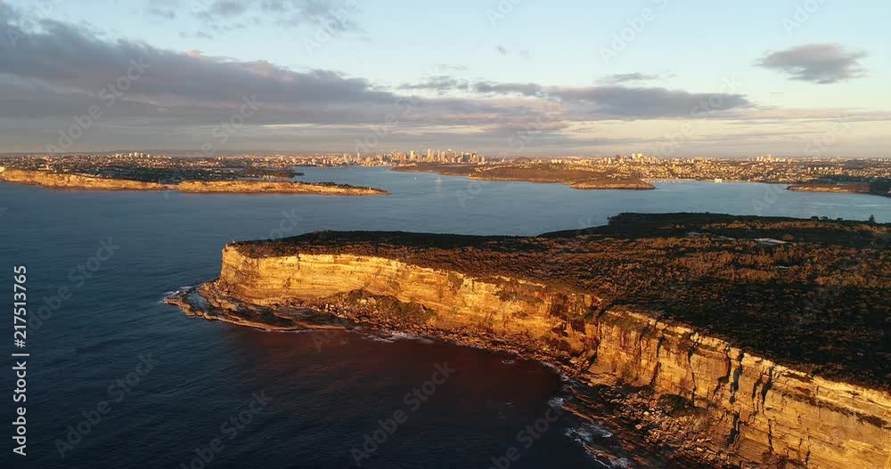 Backward flying from the edge of sandstone cliff plateau of North Head around the entrance to Sydney harbour looking at distant city CBD.
