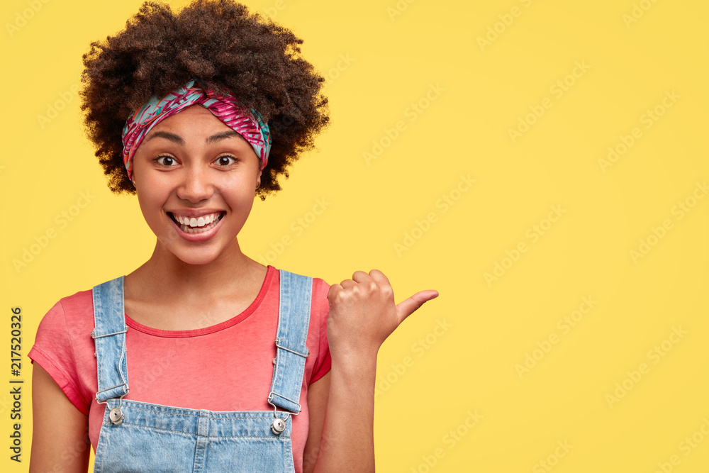 Positive young lovely female model has dark curly hair, smiles broadly, shows white teeth, indicates with thumb aside, happy to show place for entertainment, isolated over yellow studio background Positive young lovely female model has dark curly hair, smiles broadly, shows white teeth, indicates with thumb aside, happy to show place for entertainment, isolated over yellow studio background