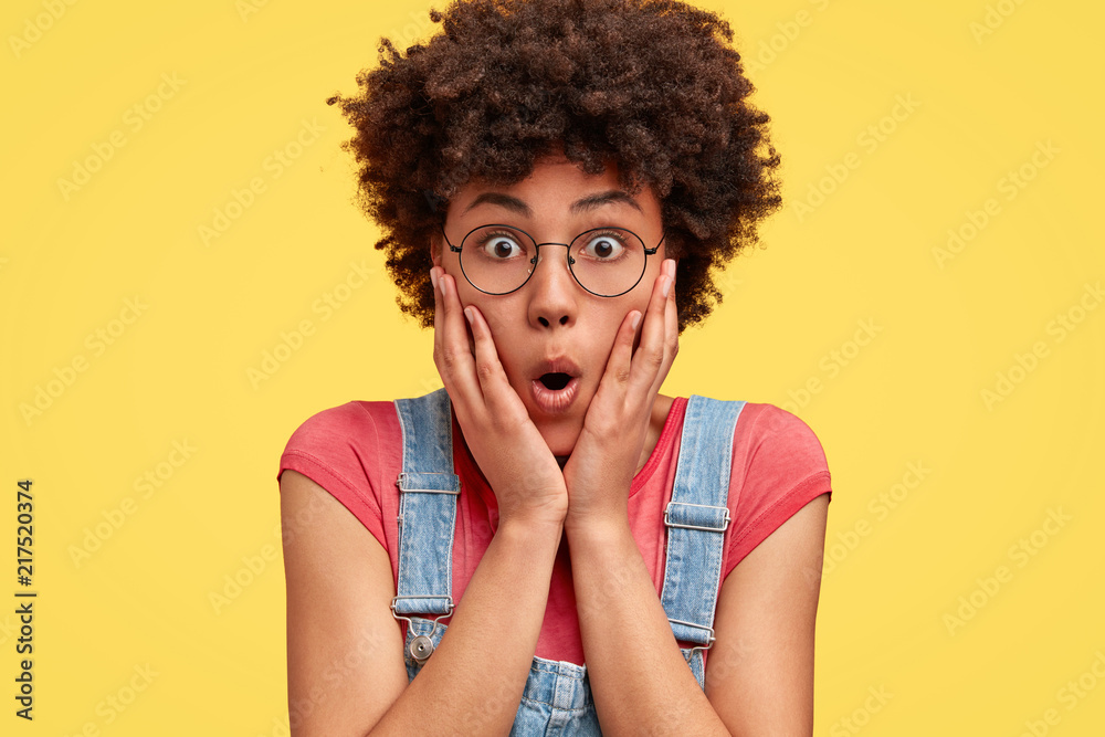Photo of surprised African American female touches cheeks, opens eyes and mouth widely, dressed in casual clothes, isolated over yellow background. Shocked mixed race woman poses alone indoor. - obrazy, fototapety, plakaty 
