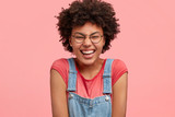Happy African American female giggles and closes eyes, shruggs shoulders, dressed in casual t shirt and denim dungarees, poses against pink background. People, positiveness, emotions concept.