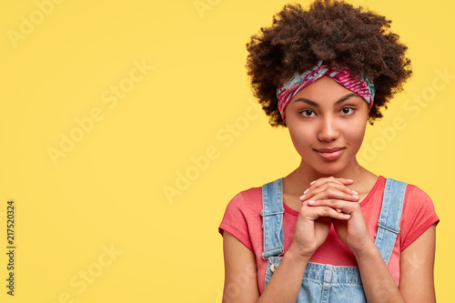 Studio shot of self confident African American woman holds hands together under chin, looks seriously directly at camera, thinks about something, stands against yellow background with copy space