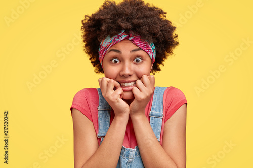 Headshot of curly female holds chin  has positive expression  opens eyes  pleasant smile  weras headband and overalls  isolated over yellow background. Charming young African American woman.