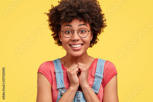 Photo of pretty happy dark skinned female with pleasant smile, keeps hands together, recieves good words of praise, has curly hair, poses against yellow background. Glad African American woman