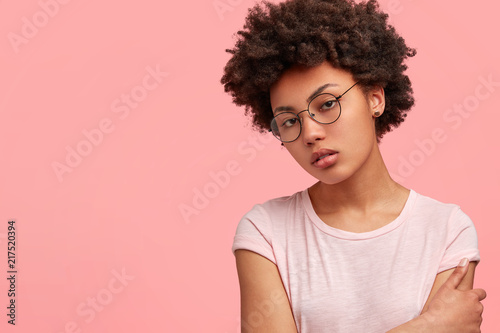 Serious dark skinned female with crisp hair, dressed in casual t shirt, has dark skin, poses against pink background with copy space for your advertisement or promotional content. Ethnicity concept