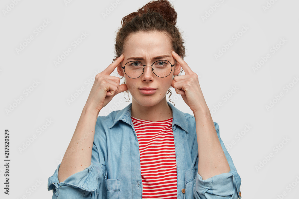 Serious blue eyed beautiful young female in spectacles, holds fingers on temples, has thoughtful clever expression, tries to remember something in mind, has freckled skin and specific appearance Serious blue eyed beautiful young female in spectacles, holds fingers on temples, has thoughtful clever expression, tries to remember something in mind, has freckled skin and specific appearance