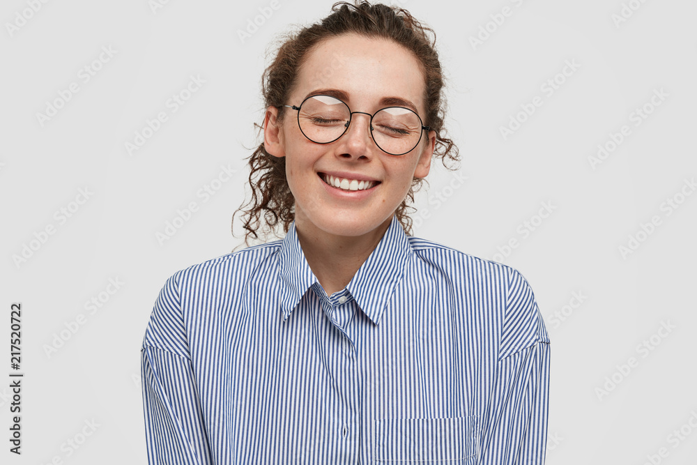 Timid young pleased freckled female closes eyes, has broad smile, imagines something fantastic, has relaxed expression, dreams about new romantic relationships, isolated over white background.