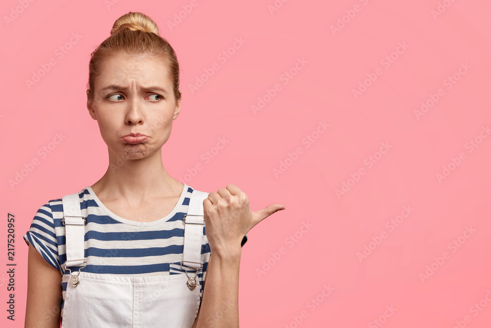 Horizontal shot of displeased gloomy blonde female purses lips and points aside with thumb, feels discontent with something, poses over pink studio wall, free space for advertisement or promotion Horizontal shot of displeased gloomy blonde female purses lips and points aside with thumb, feels discontent with something, poses over pink studio wall, free space for advertisement or promotion