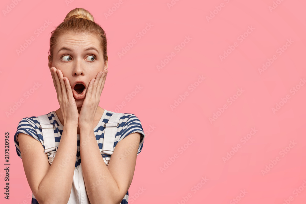 Stunned attractive female model with hair combed in bun, keeps hands on cheeks, dressed casually, notices something aside, isolated over pink background with copy space for your advertisement