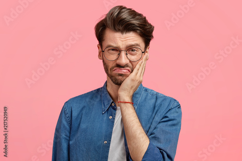 Image of displeased bearded young guy has sullen facial expression, touches cheek with hand, dressed in fashionable denim shirt, poses against pink background, being dissatisfied with something