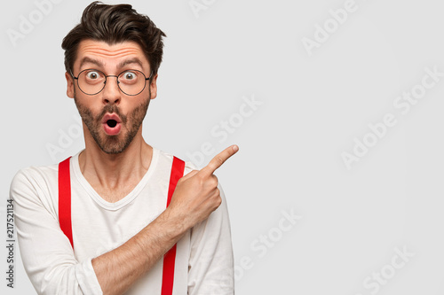 Emotional bearded male has surprised facial expression, astonished look, dressed in white shirt with red braces, points with index finger at upper right corner, shows copy space for advertisement photo