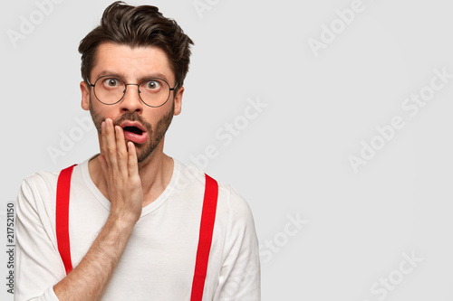 Surprised bearded man with stunned expression, keeps hand on cheek and opens mouth, has displeased face, dressed in white sweater and red suspenders, models against blank wall for your text. photo