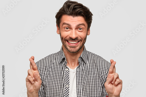 Happy Caucasian male with trendy haircut, gestures indoor, raises crossed fingers, believes in good luck, smiles sincerely, wears fashionable checkerd shirt, isolated over white wall. Expectation photo