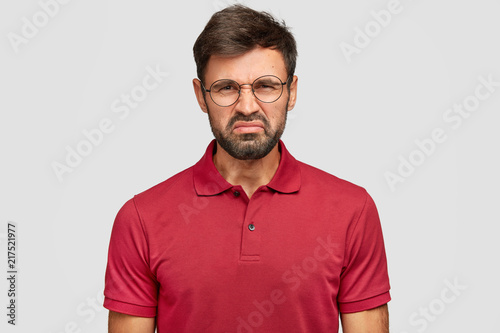 People and dislike concet. Discontent young bearded male frowns face, has dark stubble, displeased facial expression, sees something unpleasant, wears casual red t shirt, isolated over white wall photo