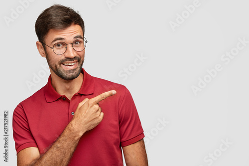 Indoor shot of positive bearded male in casual red t shirt, points with index finger aside, shows free space for your advertising content or promotional text, isolated over white background. photo