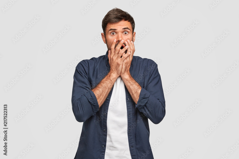 Studio shot of frightened Caucasian male covers mouth with both hands, stares with bugged eyes, dressed in fashionable outfit, reacts on something, isolated over white background. Emotions concept
