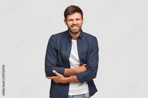 Attractive young bearded male suffers from terrible stomachache, crosses hands and clenches teeth with pain, ate some spoiled products, being ill, isolated over white background. Problems with health photo
