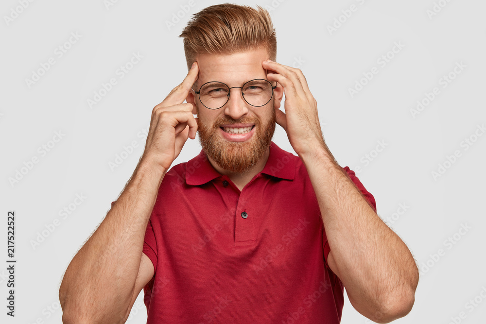 Fototapeta premium Photo of cheerful red haired young hipster has bad memory, keeps hands on temples, tries to remember something, has friendly smile, wears casual outfit, stands alone against white background.