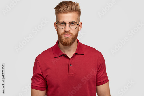 Candid shot of self confident serious Caucasian male with foxy hair and beard, dressed casually, looks directly at camera with mysterious expression, wears spectacles, isolated over white background © wayhome.studio 