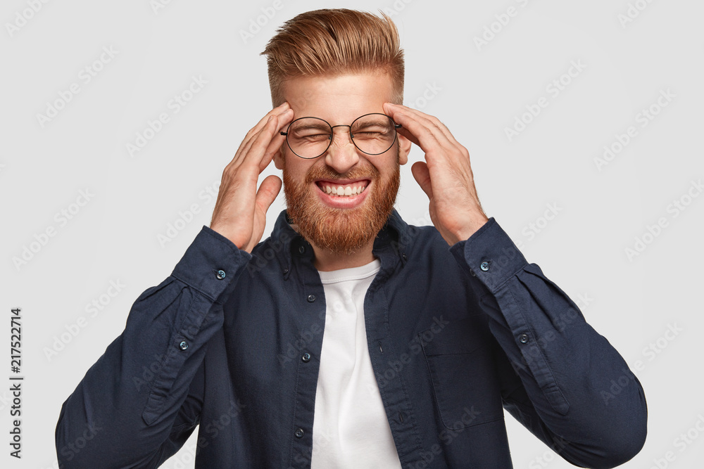 Horizontal shot of attractive stressful foxy male clenches teeth, keeps hands on temples, has headache, dressed in fashionable clothes, isolated over white background. People, emotions and feeling Horizontal shot of attractive stressful foxy male clenches teeth, keeps hands on temples, has headache, dressed in fashionable clothes, isolated over white background. People, emotions and feeling