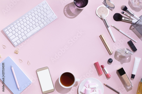 Top view of mockup of mobile phone with white blank copy space screen in female hand. Flat lay women workspace with laptop, decorative cosmetic set, stationery and flowers, hard light