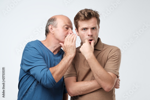 Senior man sharing secret or whispering gossips into his son ear. Telling family secret concept