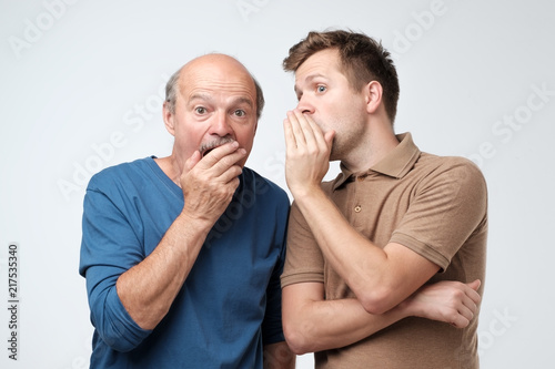 Young caucasian son telling secret to his senior father. Senior man in shock opening his mouth.
