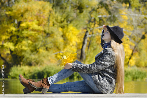 Smiling girl sitting on the pier with autumn leaves in hands. Sunny day