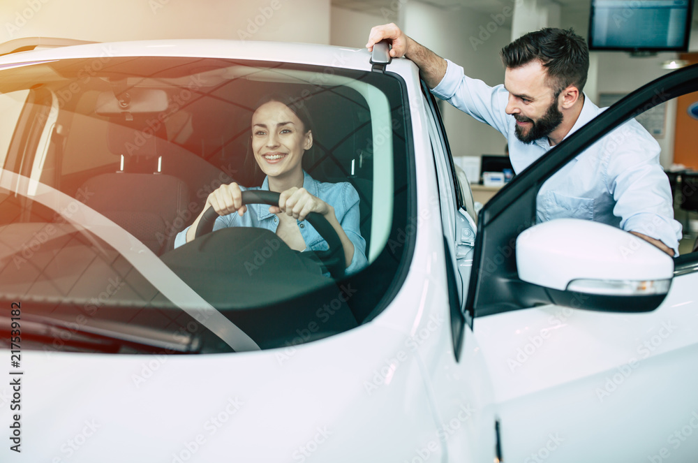 Beautiful young modern couple chooses and buying a new car for the family in the dealership. Woman inside of car tests it Beautiful young modern couple chooses and buying a new car for the family in the dealership. Woman inside of car tests it
