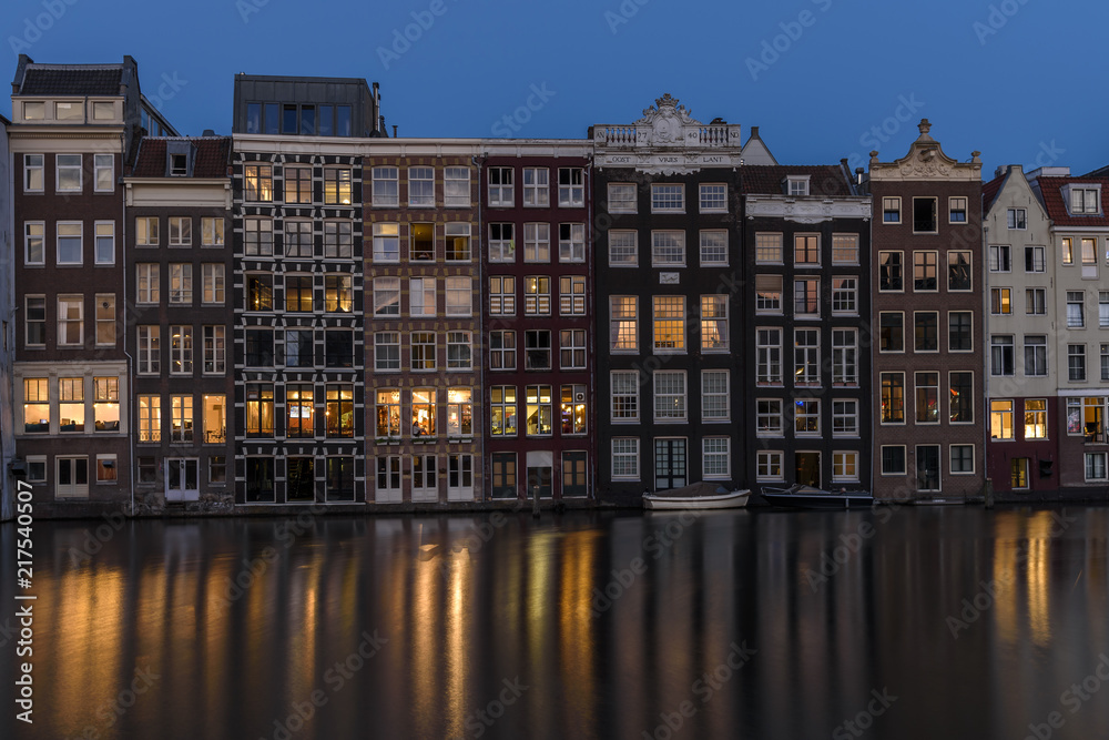 Tall buildings in Amsterdam, at dusk, with the lights from the windows reflecting on the calm water of the canal